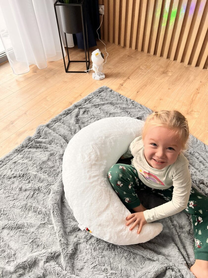 Toddler sitting in white half-moon pillow on gray rug, smiling at camera