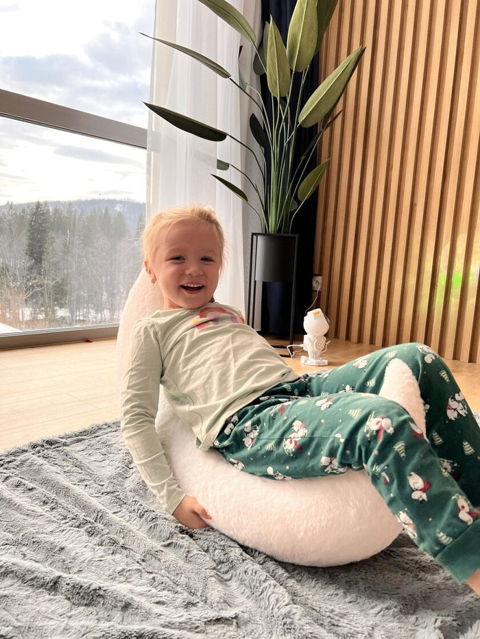 Young child sitting on white half-moon pillow by window, enjoying cozy playtime
