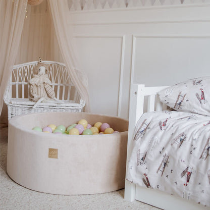 Cream fabric ball pit filled with colorful balls next to white bed