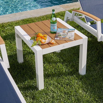 Square outdoor patio side table with white aluminum frame and slatted teak wood tabletop, poolside end table beside lounge chairs on green lawn